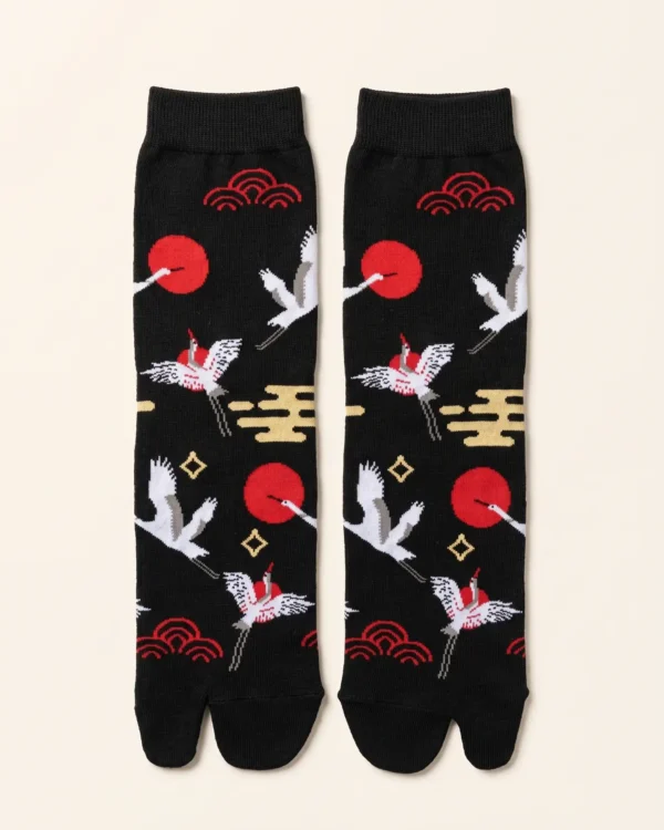 Chaussettes Tabi noires à motifs japonais (Vue à plat) : Une paire de chaussettes Tabi noires à orteils séparés, posées à plat côte à côte sur un parquet en bois clair. Elles sont décorées de motifs brodés de style japonais : des grues en vol, des vagues rouges traditionnelles et des cercles rouges (soleils). Les orteils Tabi sont clairement définis.